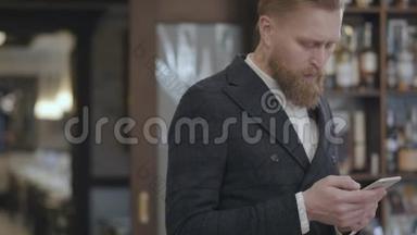 一位成功的年轻有胡子、自信的商人站在餐馆或餐馆里用手机打字的肖像